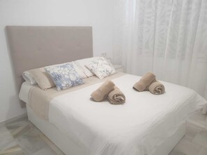 A neatly made bed with a white bedspread and two rolled towels, accompanied by floral-patterned pillows.