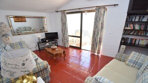 Ein Wohnzimmer mit einer Couch, einem Couchtisch, einem Fernseher, einem Bücherregal und einem großen Fenster mit Vorhängen.