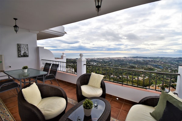 Uma varanda com mobiliário de vime, uma mesa de vidro e vista para a cidade.