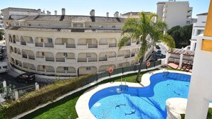 A multi-story building with a swimming pool, a palm tree, and a road with a car. - Pueblo Torrequebrada, Benalmadena 2 Bed Apt (Benalmádena)