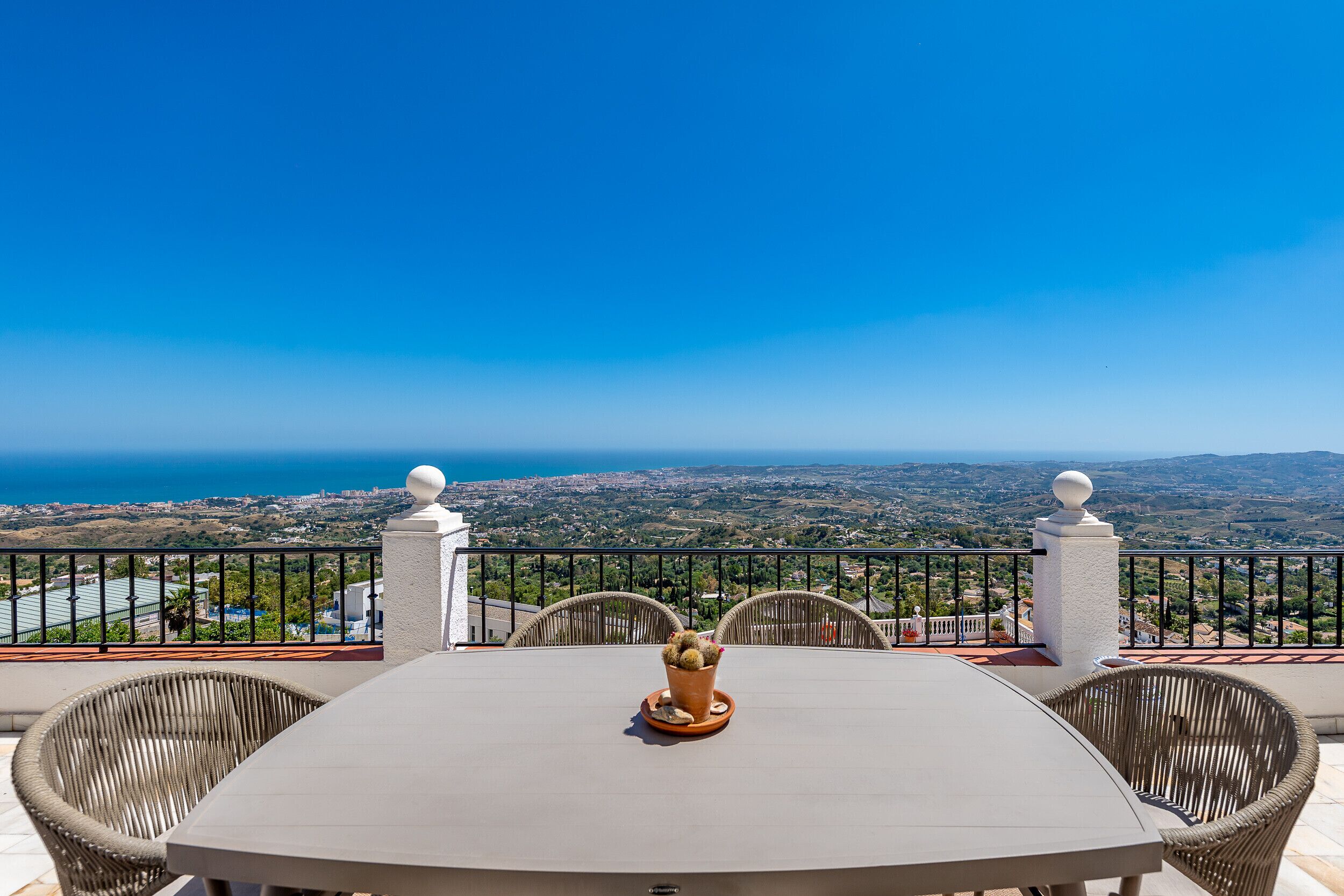 A balcony with a table and chairs, a potted plant, and a view of the sea and landscape.