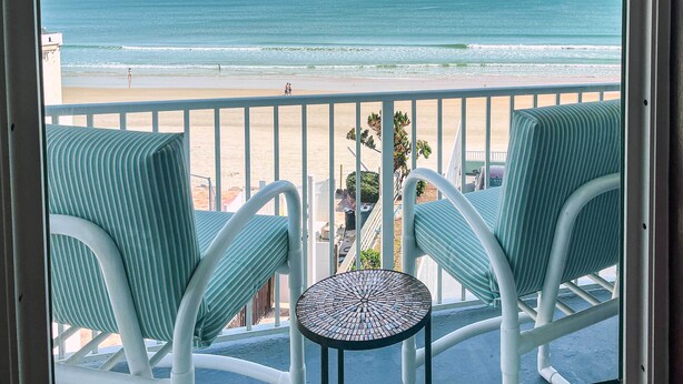 A balcony with two striped chairs and a small table, overlooking a beach with people and palm trees. - Oceanfront Daytona Beach Condo w/ New Pool! (Daytona Beach)
