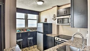 A compact kitchen with granite countertops, a stainless steel refrigerator, a microwave, and a stove. There are two bar stools and a window with a view. - Oceanfront Daytona Beach Condo w/ New Pool! (Daytona Beach)