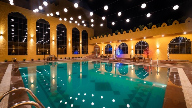 Una piscina con superficie de agua cristalina, rodeada por un edificio con amplias ventanas arqueadas y luces colgantes.