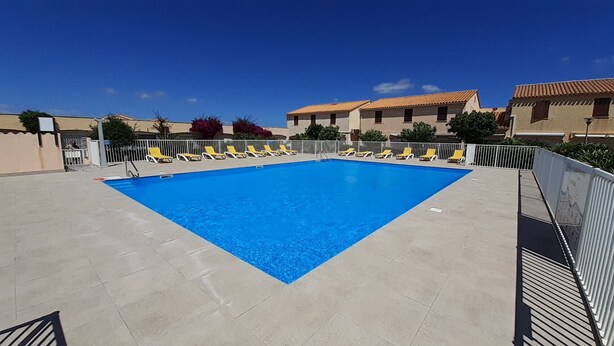Piscina retangular com deck ladrilhado, cercada por uma vedação branca e cadeiras de descanso amarelas.