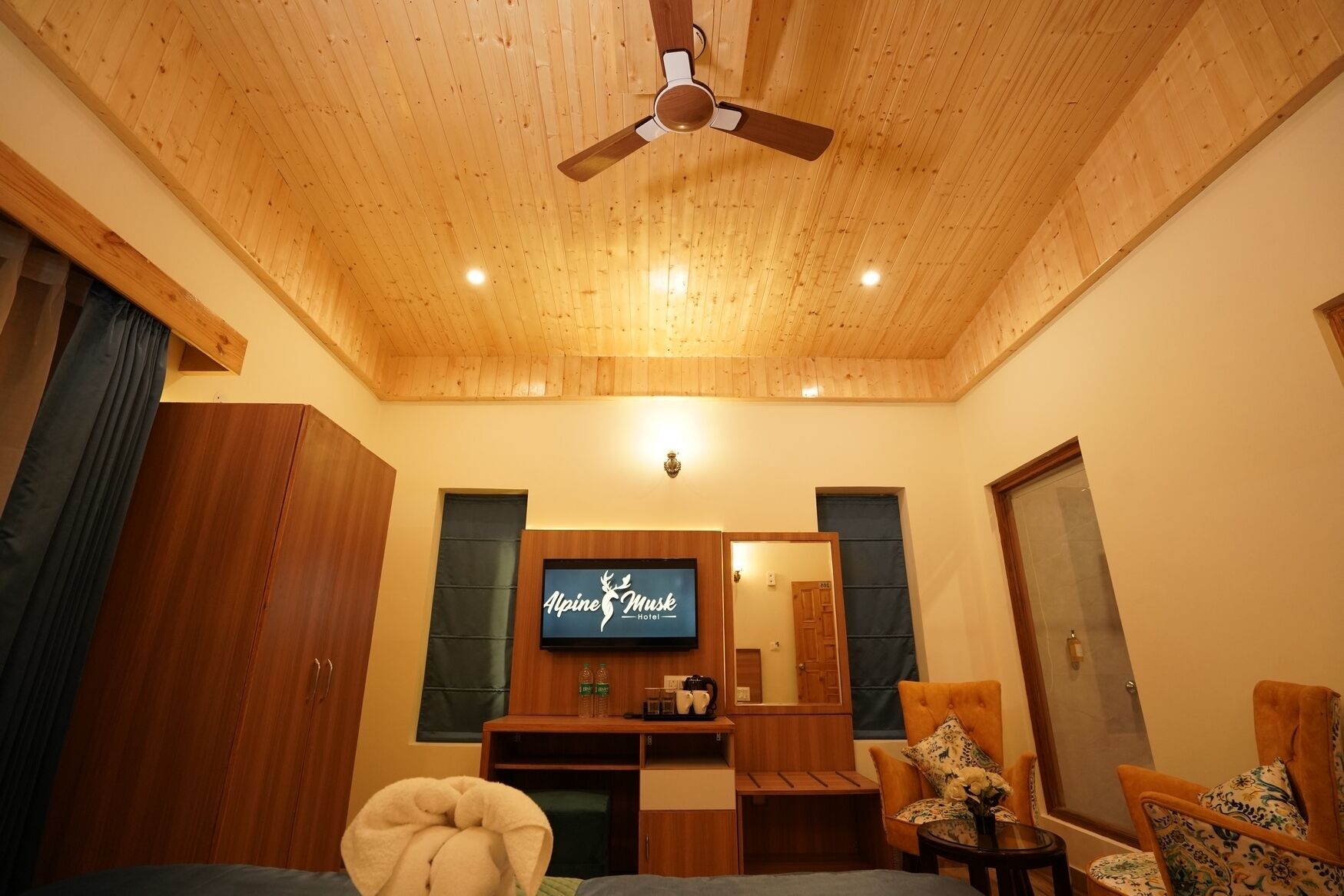 A cozy hotel room with wooden ceiling and walls, a television mounted on the wall, a wooden cabinet, and a comfortable seating area with a chair and a small table.