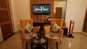 A hotel room with two armchairs, a small table with wine glasses, a television displaying 'Alpine Musk Hotel', and a standing heater. - HOTEL ALPINE MUSK (Tehri)