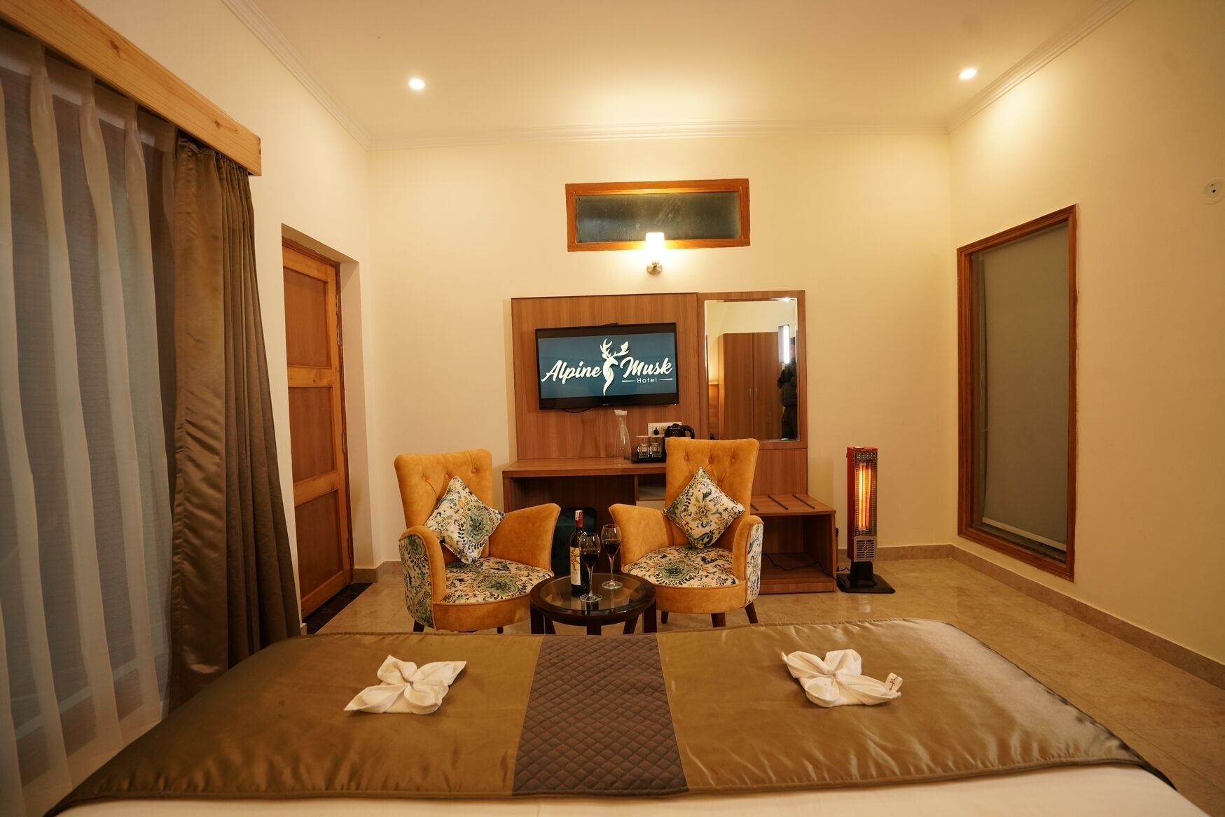 A hotel room with two armchairs, a small table with a bottle of wine, a flat-screen TV, and a bed with a brown blanket.