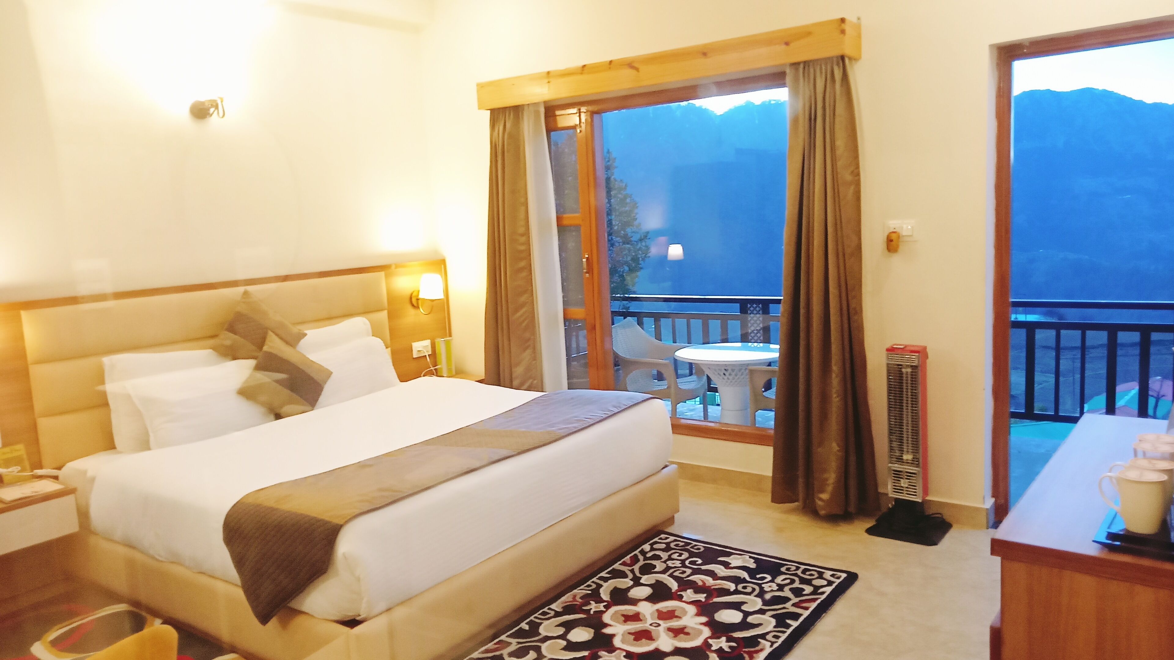 A hotel room with a bed, a nightstand, a table with a chair, and a view of mountains through the balcony.