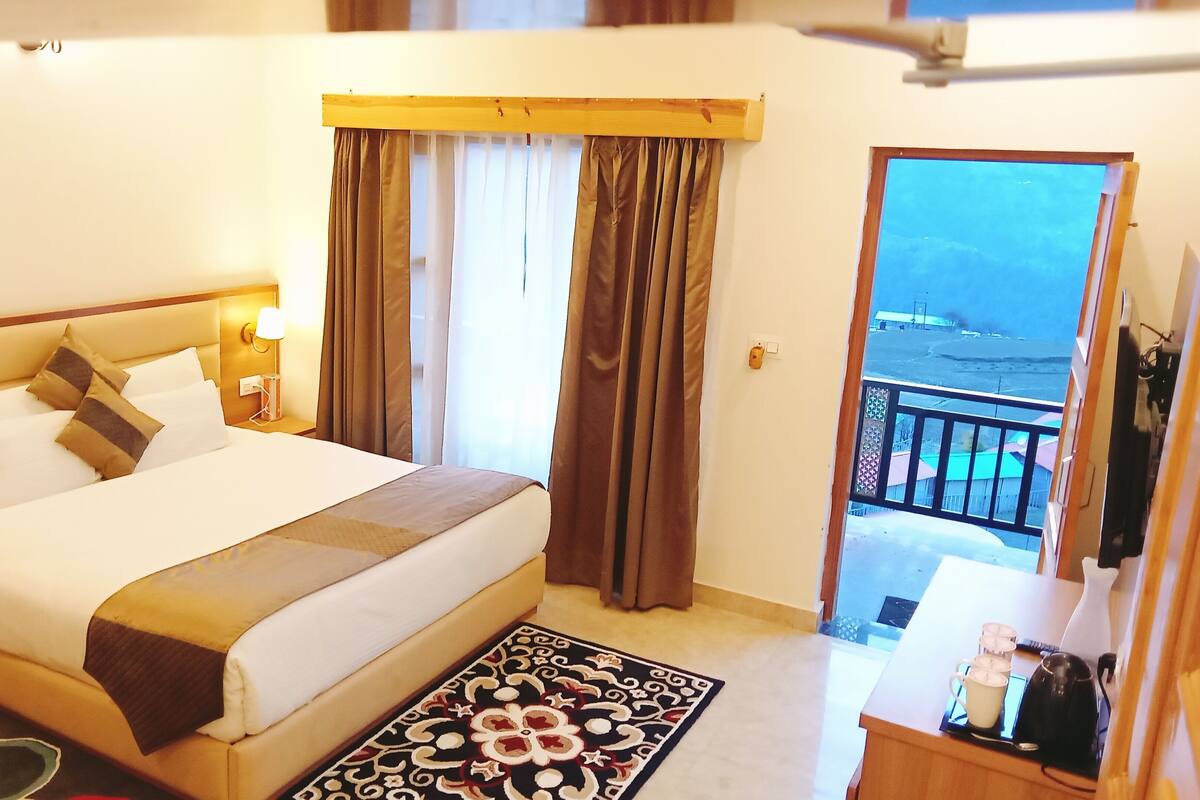 A hotel room with a bed, a nightstand, a lamp, a window with curtains, a balcony with a view, and a small table with a kettle and cups.