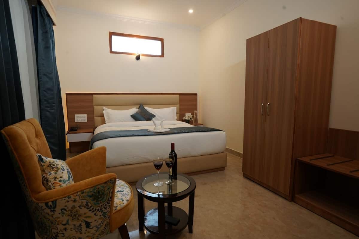 A hotel room with a bed, a wooden wardrobe, a small table with a wine bottle, and a patterned armchair.