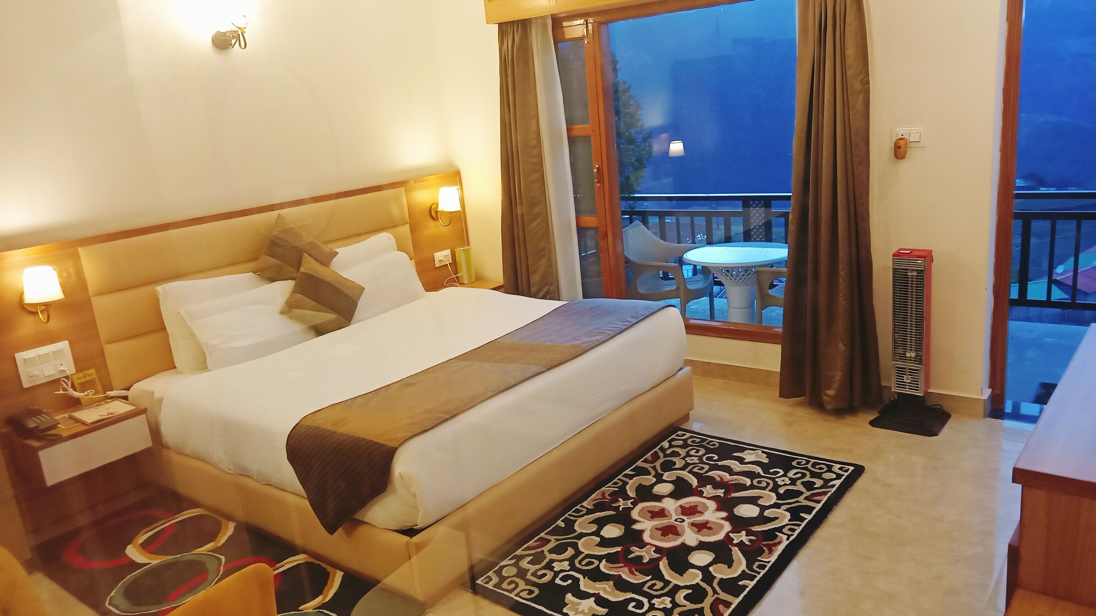 Habitación de hotel con una cama grande, lámparas de noche, una mesita y vistas al exterior.