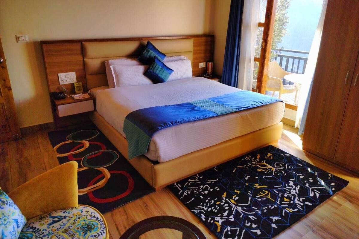 A well-lit hotel room with a large bed, a wooden headboard, a nightstand, a chair, and a view of the outdoors through a window.