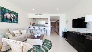 A modern living room with a sofa, a coffee table, and a TV. There is a kitchen area with a dining set and a large window.
