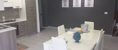 Una cocina moderna con armarios blancos, una estufa a gas y un comedor con una mesa y sillas blancas. Hay tres cuadros en la pared.