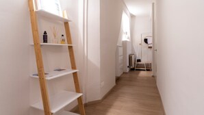 A narrow hallway with white walls and wooden flooring. There are white shelves with various items on them, including a bottle and a vase. A white chair is visible in the distance. - Italianway - San Vitale 87 (Bologna)
