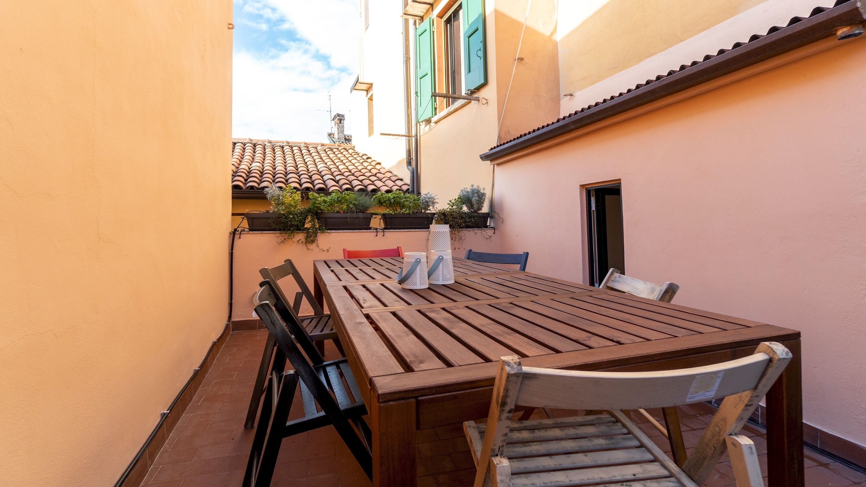 A wooden table and chairs in a narrow outdoor space with a tiled roof and plants.