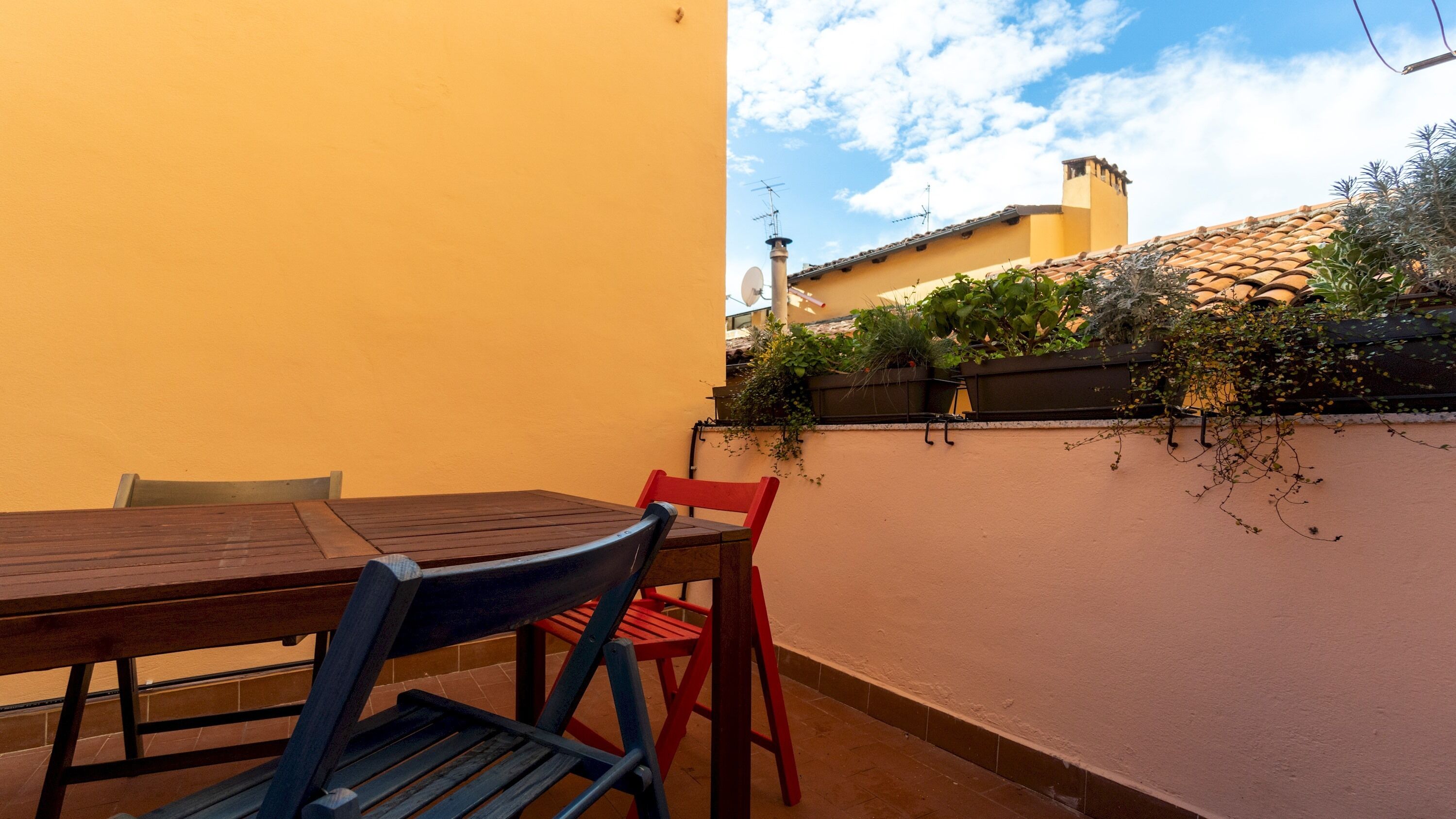 Une terrasse avec une table et des chaises en bois, un mur jaune et une vue sur un bâtiment doté d’un toit en tuiles.