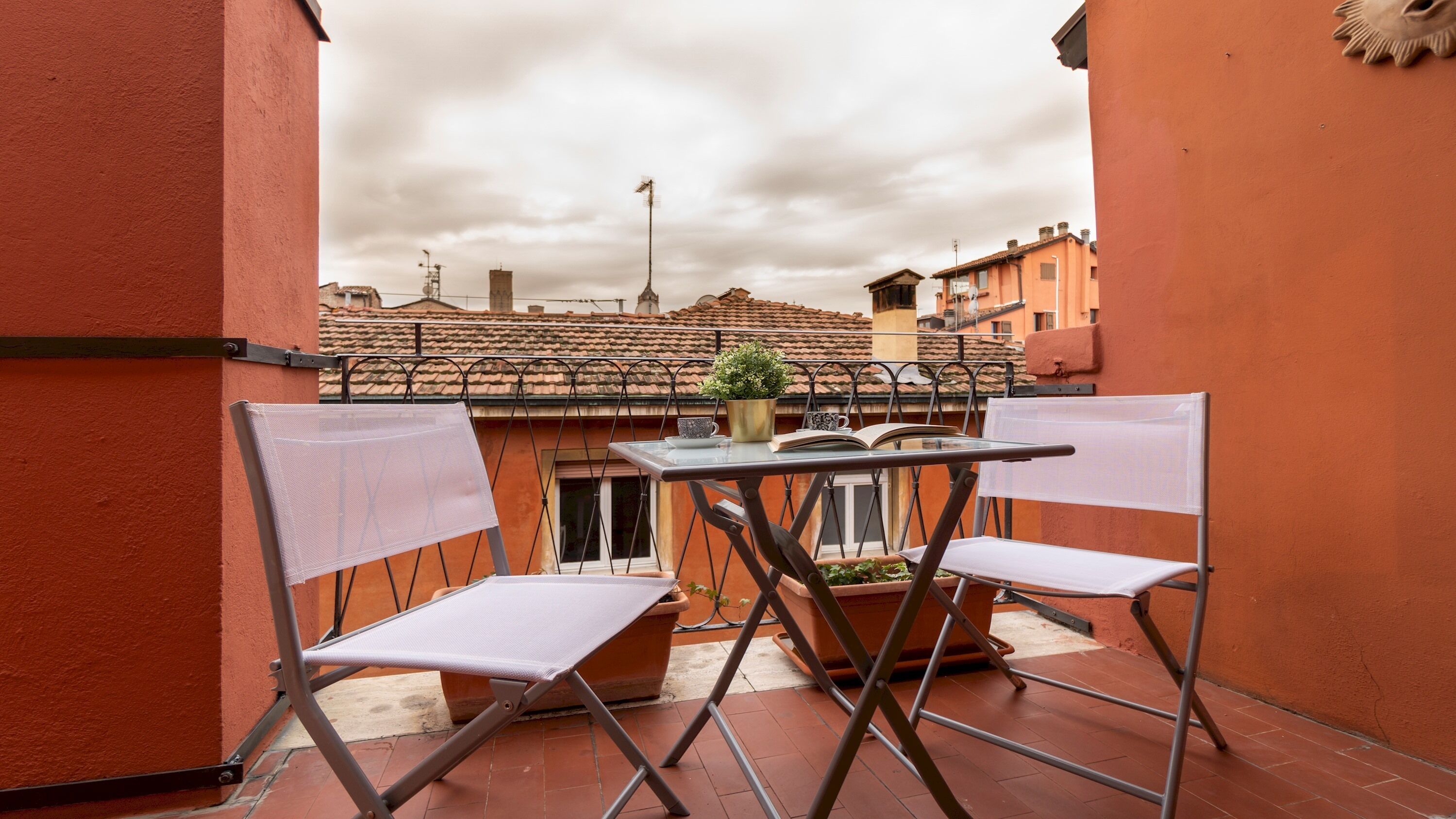 Une petite terrasse extérieure avec une table et deux chaises, un pot de fleurs et une vue sur les bâtiments voisins.