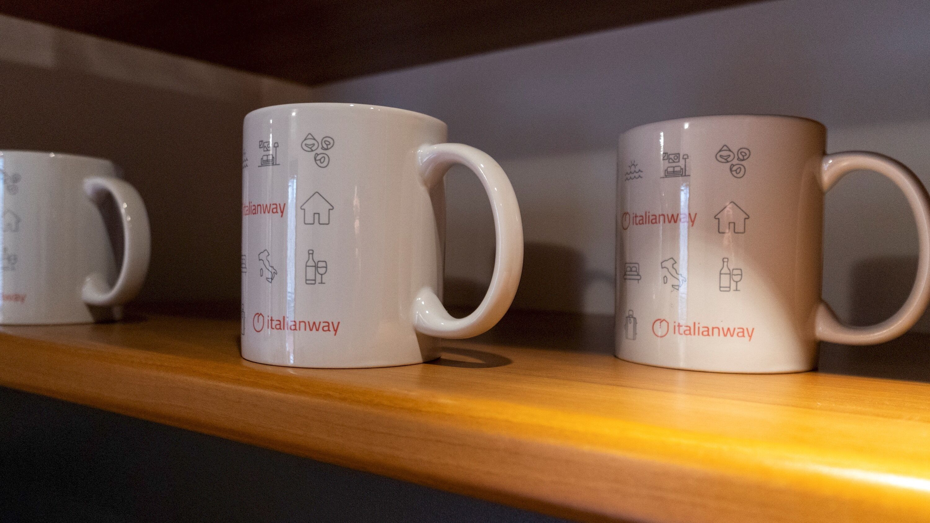 Three white ceramic mugs with Italian symbols and the text "italianway" on a wooden shelf.