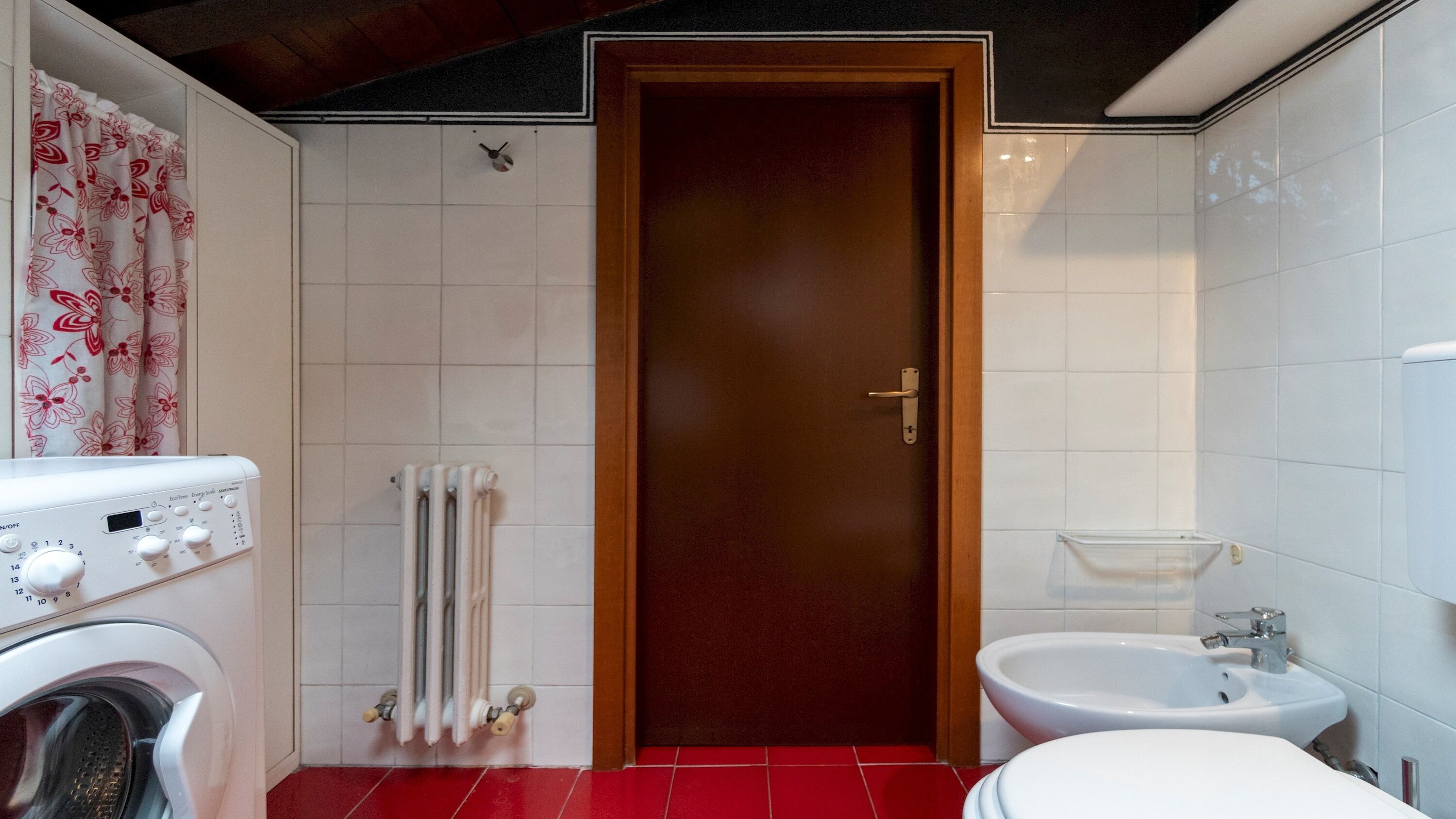 A compact bathroom with a wooden door, a washing machine, a radiator, and a bidet.