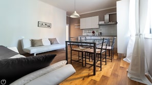 A modern living room with a glass dining table, a sofa, and a kitchen in the background.