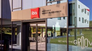 Una entrada de hotel moderna con fachada de vidrio, un logotipo rojo y un cartel que dice "IBIS BRUSSELS GROOT-BIJGAARDEN".