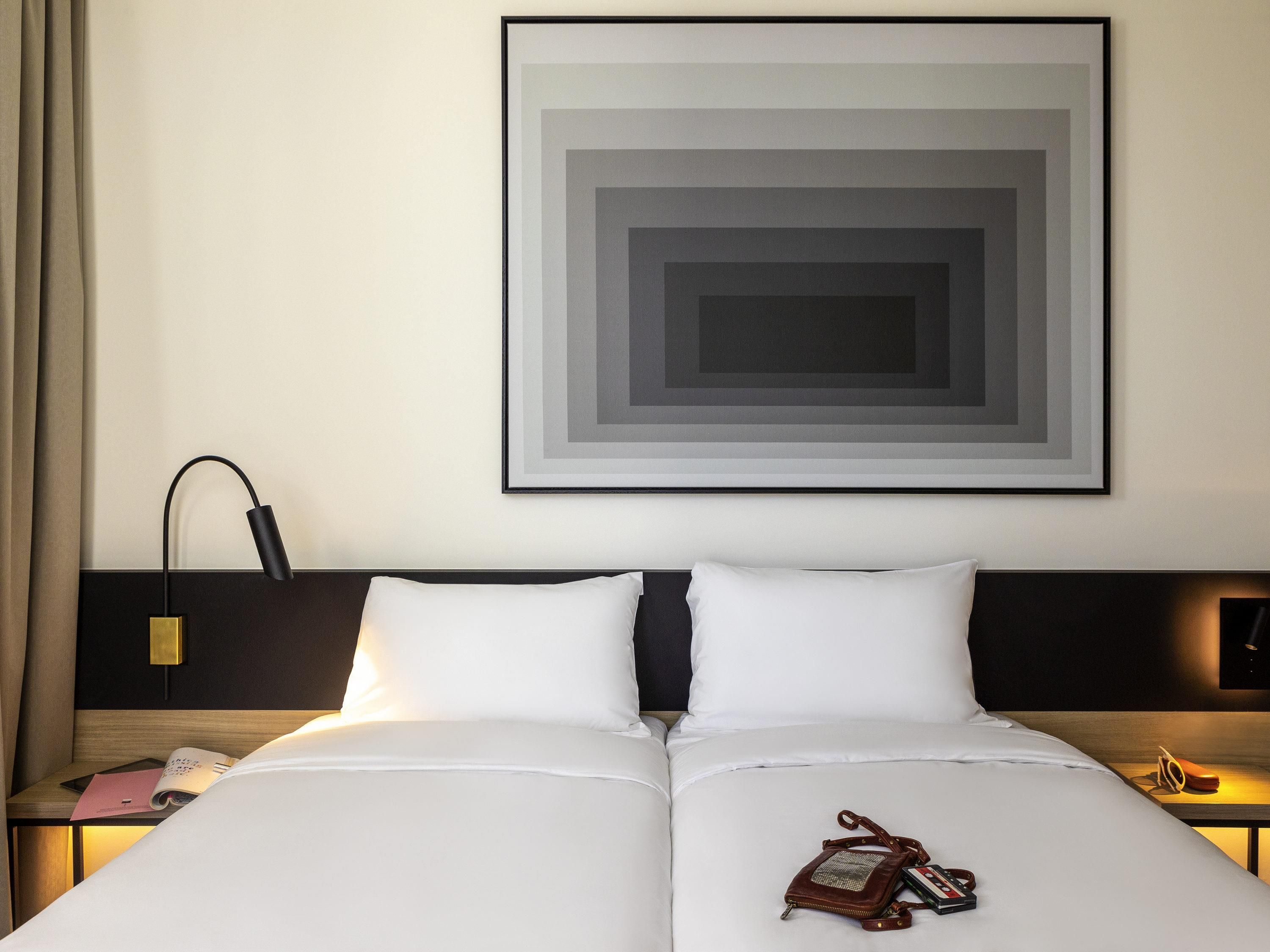 A hotel room with a bed, a nightstand, a lamp, a framed artwork, and a handbag on the bed.