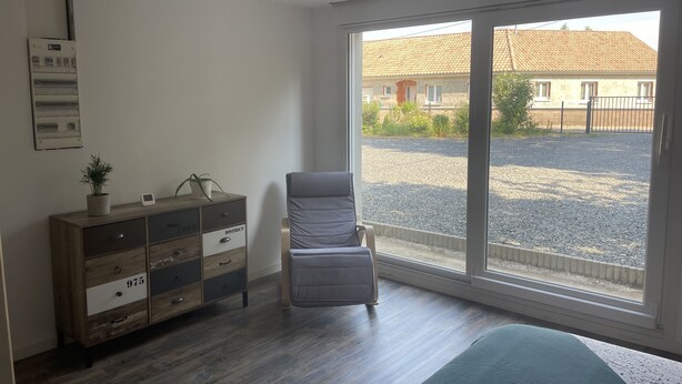 Une pièce avec une grande fenêtre, une chaise, une commode en bois et un lit.