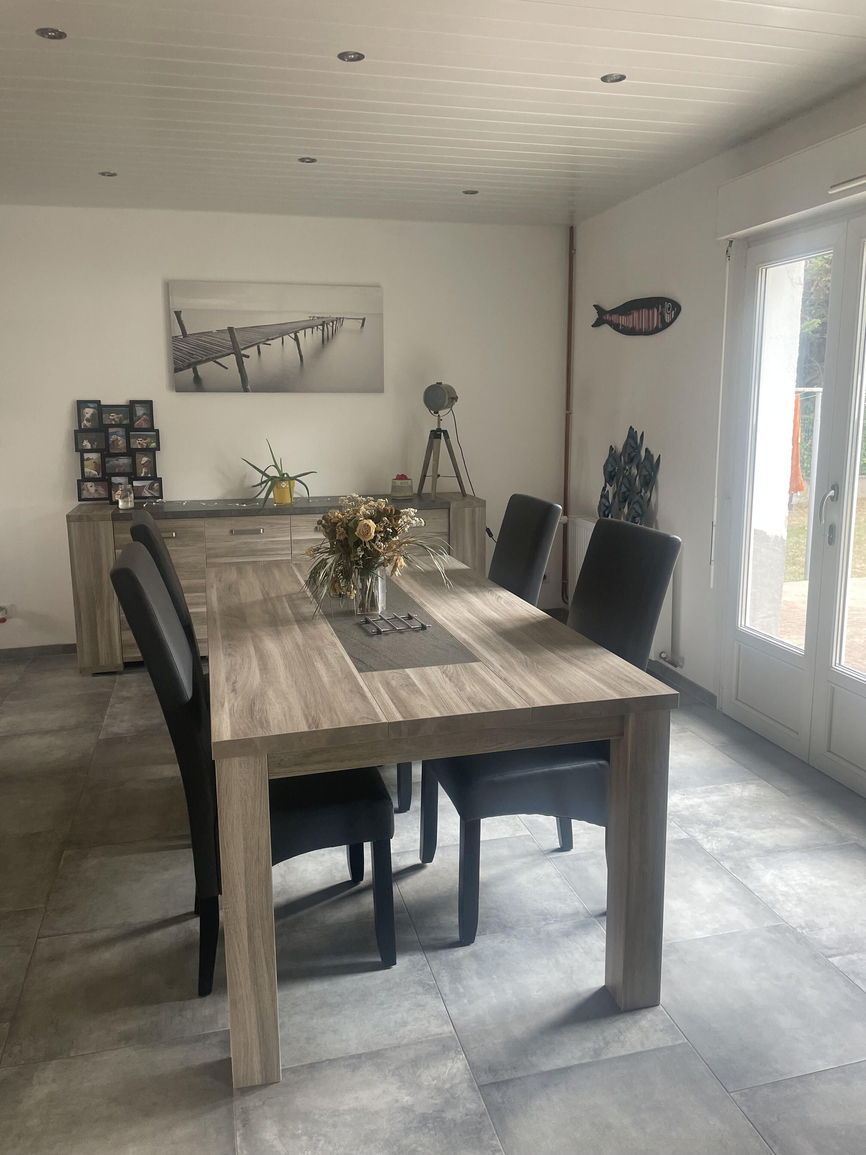Un coin repas avec une longue table en bois, des chaises noires, un vase de fleurs, une décoration en forme de poisson et un tableau encadré au mur.