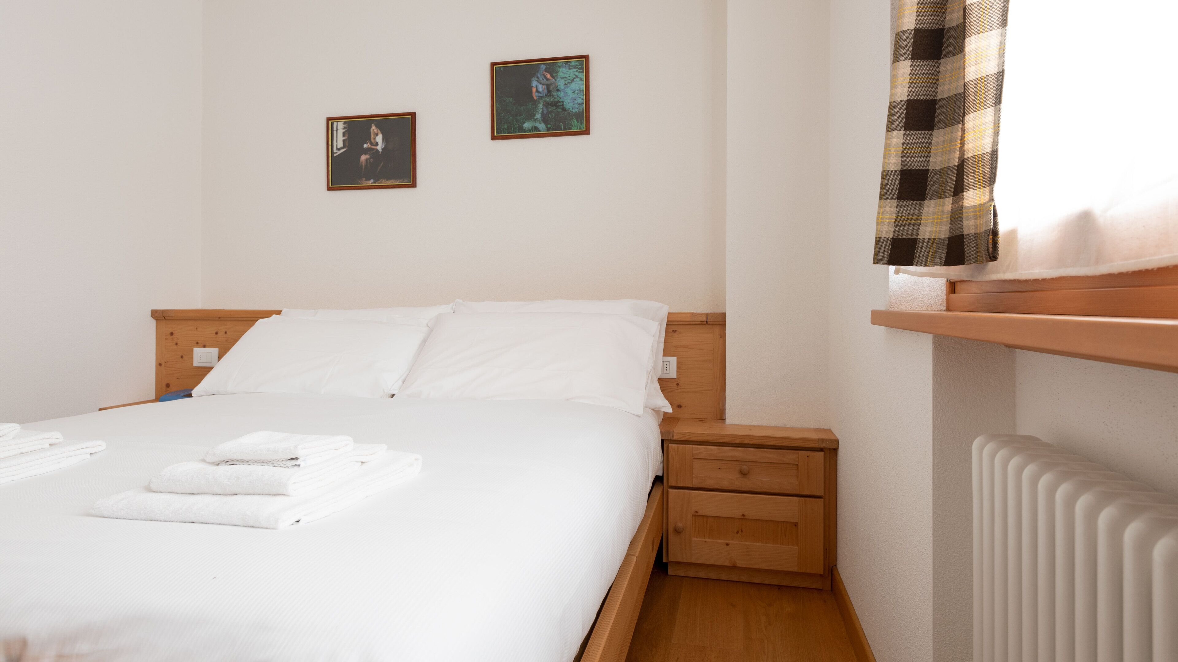 A neatly made bed with white linens and towels, a wooden headboard, a nightstand with a drawer, and a radiator. Two framed pictures are hung on the wall.