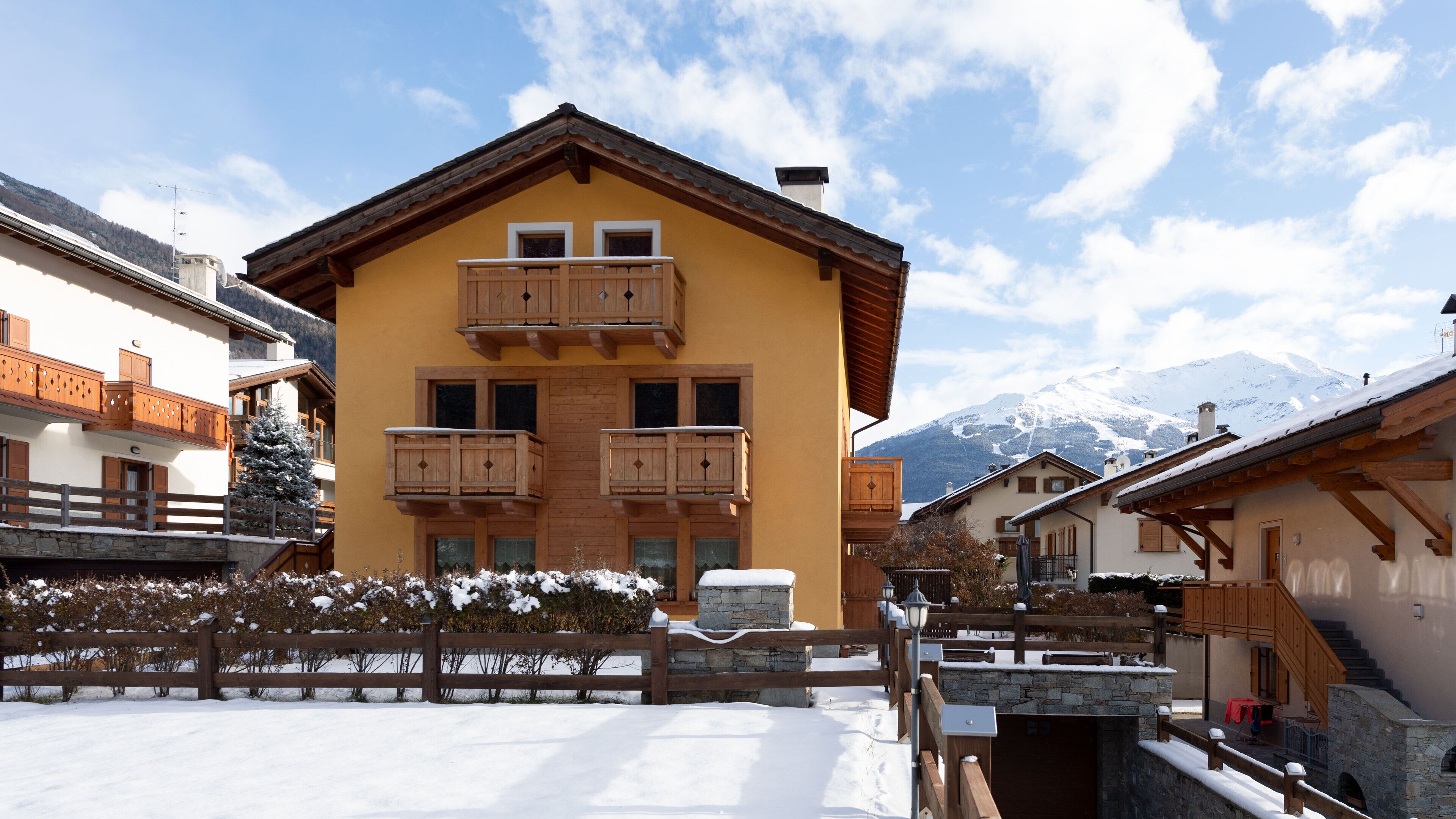 Un bâtiment jaune avec des balcons en bois, une entrée en pierre et une montagne enneigée en arrière-plan.