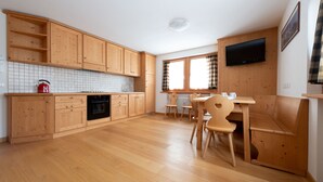 A kitchen with wooden cabinets, a red kettle, a black oven, and a dining area with wooden chairs and a table. - Adda 10 (Valdidentro)