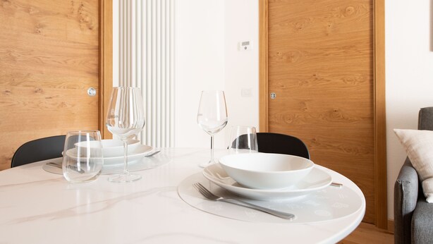A dining table set with white plates, bowls, and wine glasses, with a chair and a sofa in the background.