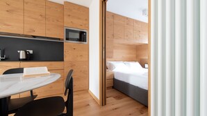 A modern hotel room with a wooden interior, a dining area with a table and chairs, a microwave, and a bed with a gray mattress.