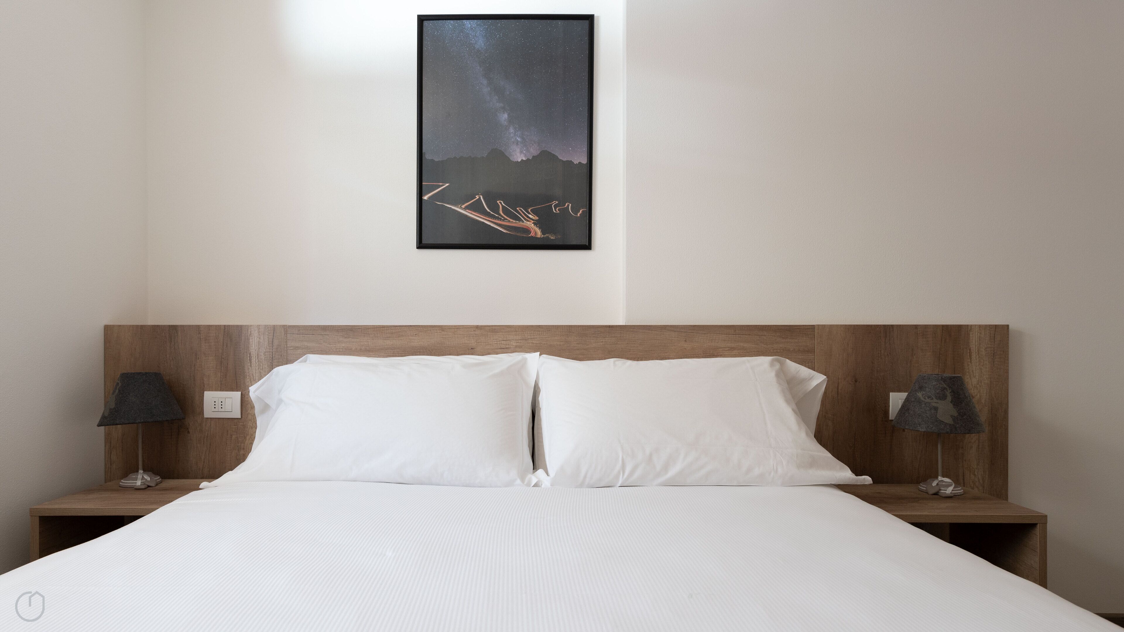A hotel room with a large bed, two bedside tables with lamps, a framed picture of a mountain and stars on the wall, and a wooden headboard.