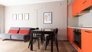 Ein modernes Wohnzimmer mit einem grauen Sofa, roten Kissen, einem schwarzen Esstisch und Stühlen. Es gibt eine Küche mit orangenen Schränken und einer weißen Arbeitsplatte.