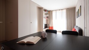 Ein modernes, helles Zimmer mit einem dunklen Holztisch, einem offenen Buch und zwei Tassen. Ein Bücherregal mit Büchern und Dekorationselementen, ein schwarzer Sessel und eine Couch mit roten Kissen sind ebenfalls vorhanden.