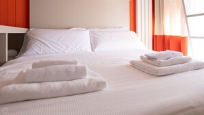 Ein ordentlich bezogenes Bett mit weißen Leinen und gefalteten Handtüchern, eine schlichte Wand und orangefarbene Vorhänge.