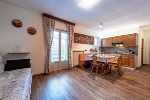 A cozy room with a bed, a dining table with chairs, a kitchen area with wooden cabinets, and a window with curtains.
