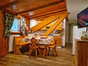 Interior acolhedor de uma cabana de madeira, com área de cozinha, armários de madeira, mesa de jantar com cadeiras e uma televisão fixada na parede.