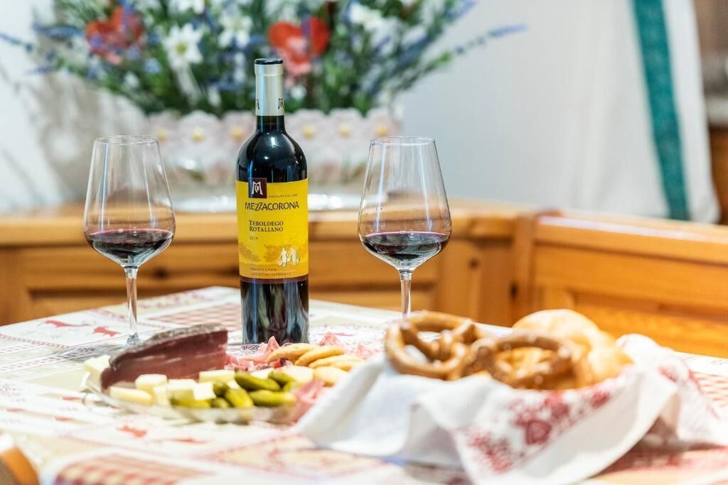 Une bouteille de vin Teroldego Rotallano, deux verres de vin rouge et une assiette avec du fromage et des bretzels, le tout posé sur une table.