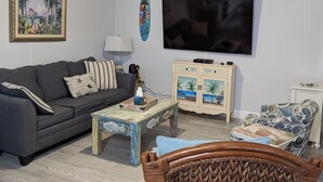 Uma sala de estar com um sofá cinza escuro, uma mesa de centro de madeira, uma cadeira de vime, uma televisão e um armário com uma cena de praia.