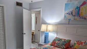 Quarto com colcha acolchoada florida, ventoinha no teto, luminária na parede e uma pintura colorida sobre a cama.