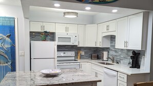 Uma cozinha moderna com armários brancos, bancada de granito e micro-ondas. Há uma área de jantar com banco e cadeiras, e um objeto decorativo na parede.