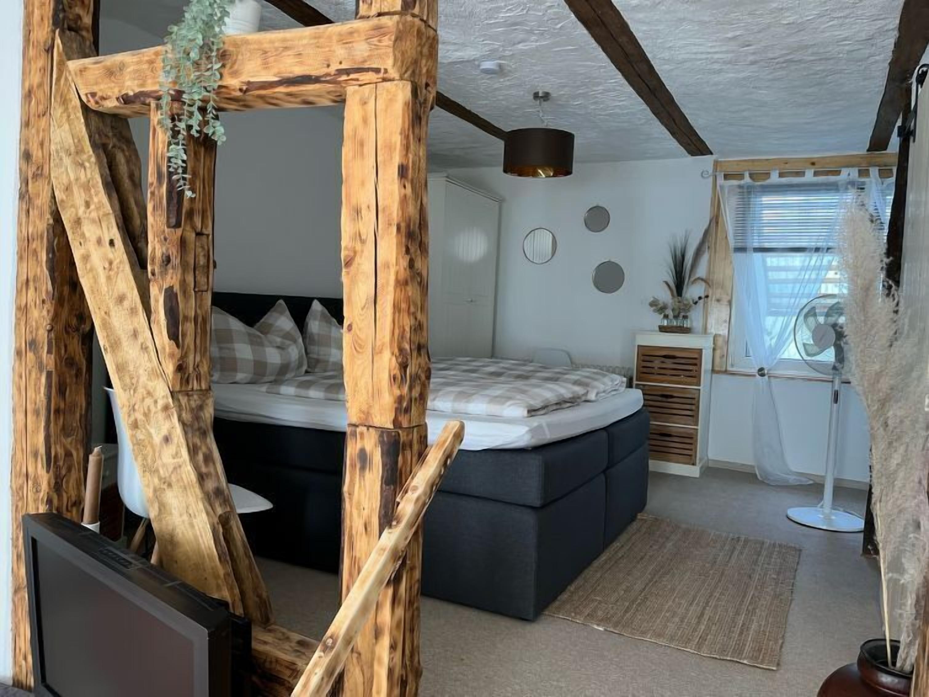 A cozy bedroom with a large bed, wooden beams, a ceiling fan, and a window with blinds.