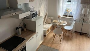 A compact kitchen with a microwave, stove, and cabinets. There is a dining area with a table and chairs, a window with curtains, and a lamp.