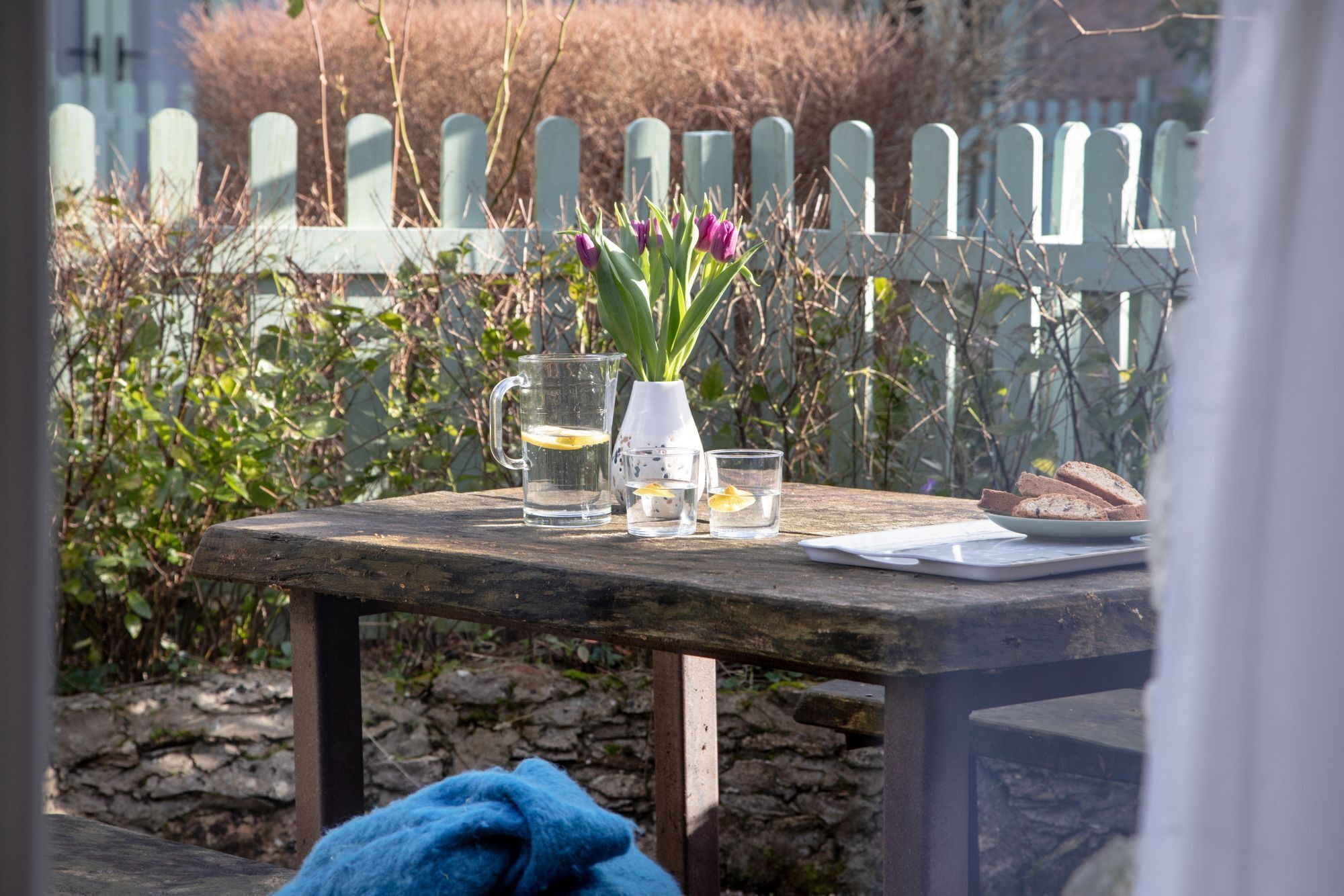 Une table en bois, sur laquelle se trouvent un pichet en verre, deux verres, une assiette avec du pain et un vase rempli de fleurs.