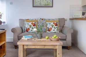 A cozy living room with a grey sofa, two patterned pillows, a wooden coffee table with a book, a vase, and a basket of green apples.