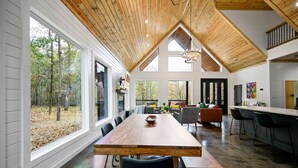Un'ampia sala da pranzo con un lungo tavolo in legno e sedie, ampie finestre che danno vista sul bosco e un soffitto alto con travi a vista.